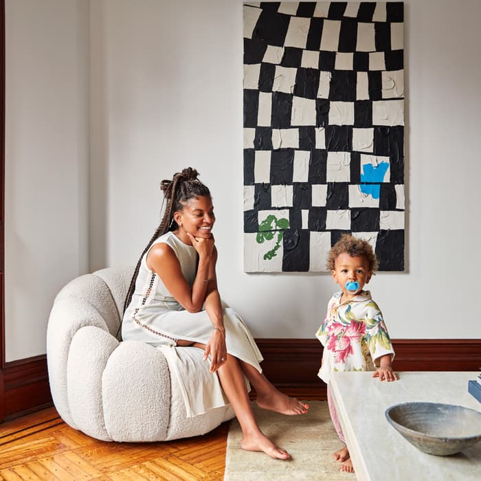 A woman in a white dress sits on a round chair, smiling at a toddler with a pacifier standing beside a white table, in an art-adorned room with large windows and wooden floor.