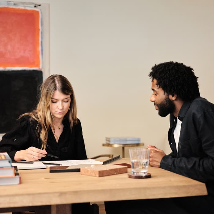 Two individuals sit at a wooden table with books and a glass of water; one person points at papers while the other attentively listens. A colorful abstract painting hangs on the wall behind them.