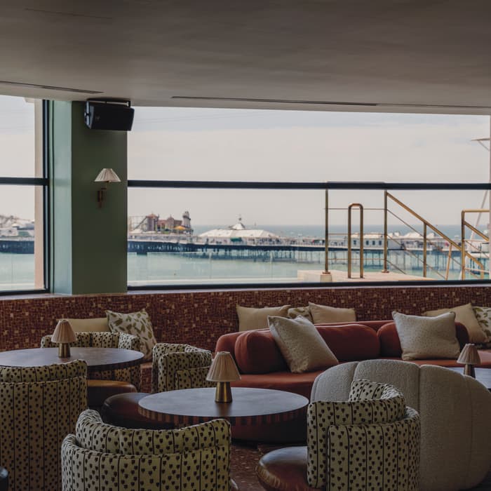 Round tables and cushioned seating face large windows overlooking a seaside pier. The interior features patterned armchairs, sofas, and small lamp fixtures, with views of the ocean and pier structures.