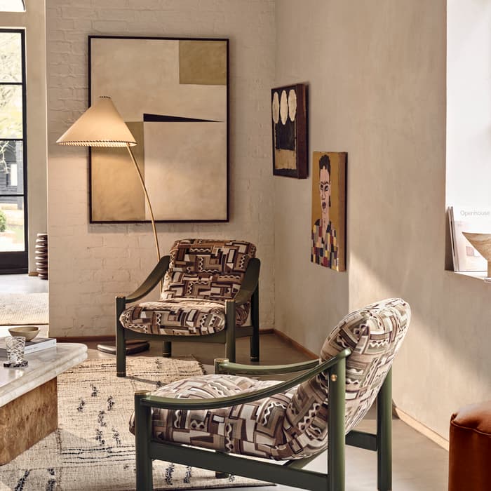 Two patterned armchairs sit in a cozy living room with modern art on the walls. A large window overlooks greenery, and a marble coffee table holds books and a glass.