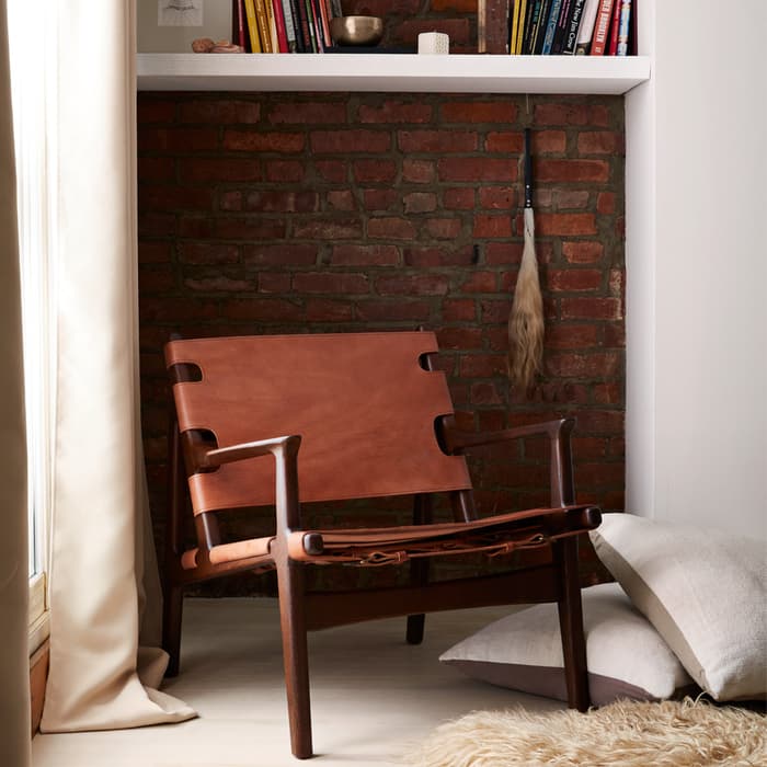 A leather chair sits in a cozy corner with a brick wall, near a window with curtains, alongside cushions on the floor and a bookshelf above containing various books and decor items.