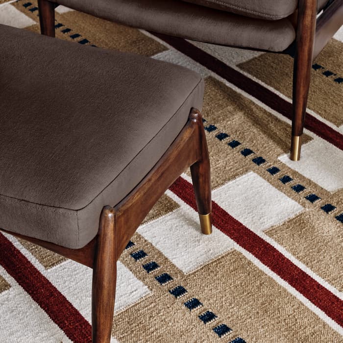 A dark brown cushioned ottoman and chair with wooden legs capped in gold, placed on a geometric-patterned rug with red, beige, white, and blue accents.