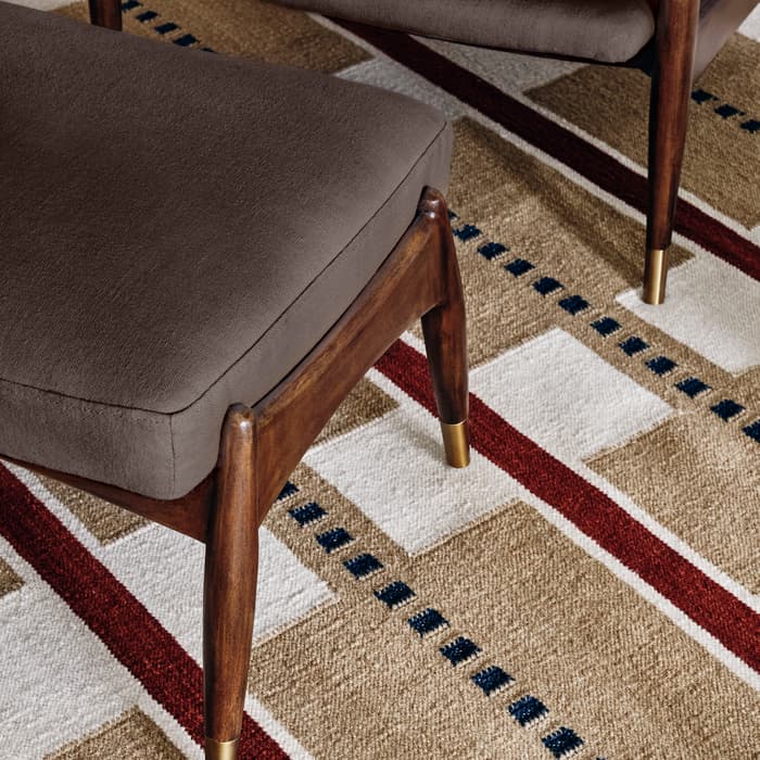Dark brown cushioned chair and matching ottoman with wooden legs and brass tips, placed on a patterned, multicolored woven rug featuring beige, red, white, and dark blue geometric designs.