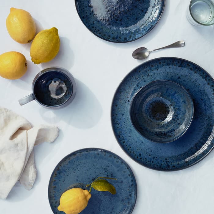Blue ceramic plates and a mug are placed on a white tablecloth. Three lemons are arranged beside the ceramics, and a silver spoon is next to a clear glass of water.