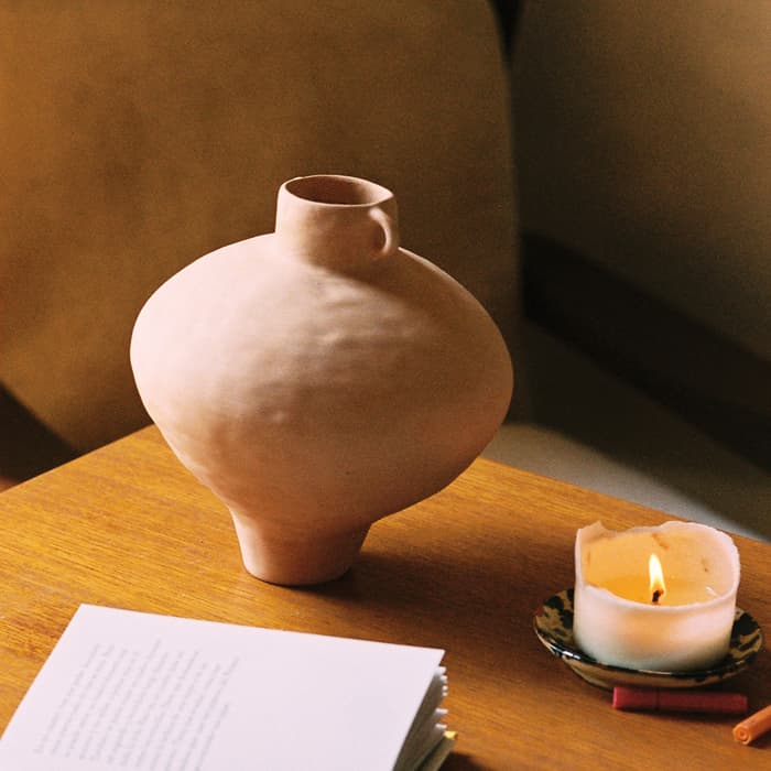 Clay vase sits on a wooden table beside an open book and a lit candle. Background features a beige armchair in a softly lit room.