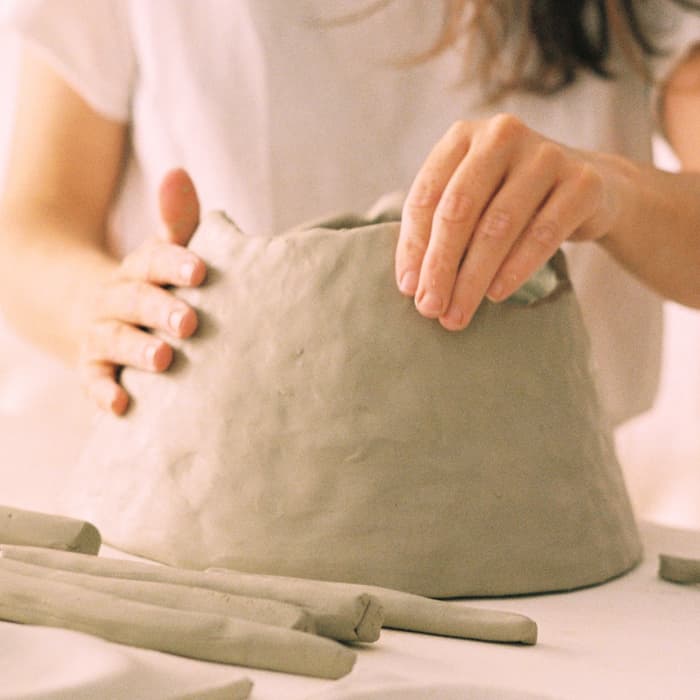 A person in a white shirt sculpts a clay object on a table, surrounded by clay tools and flat clay pieces, in an indoor setting.