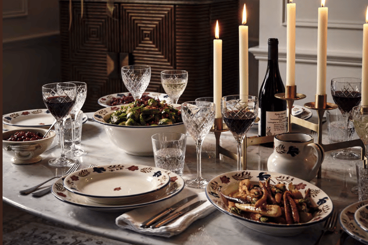 A dining table is set with plates, crystal glasses, and various dishes, surrounded by lit candles in a cozy, dimly lit room, showcasing a cabinet and decorative items in the background.