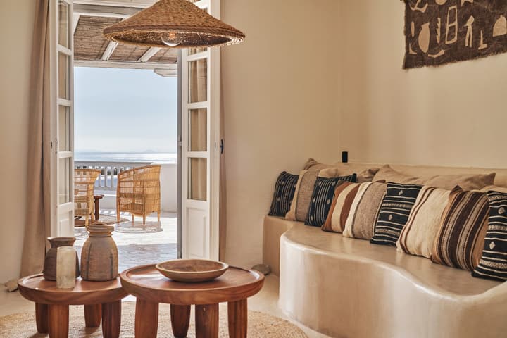 Wooden coffee tables with ceramic vases are set in a cozy, pillow-filled living room which opens onto a sunlit terrace featuring wicker furniture and an ocean view in the background.