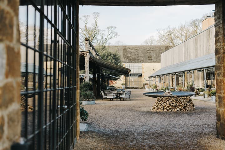Outdoor courtyard features metal chairs around tables, surrounded by rustic brick and glass buildings with large windows, shaded awnings, and greenery, with a central stone fountain or fire pit on a woodpile.