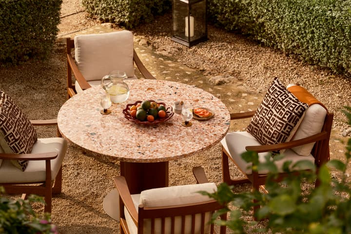 A round, stone table set for a casual beverage with a pitcher of lemon water, glasses, fruit bowl, and small plates; surrounded by cushioned chairs with patterned pillows, in a garden.