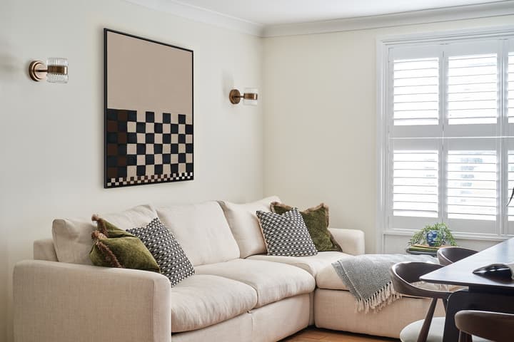 A beige sectional sofa adorned with patterned cushions sits in a bright living room, featuring wall sconces, a checkered abstract painting, and white shutters on a window.