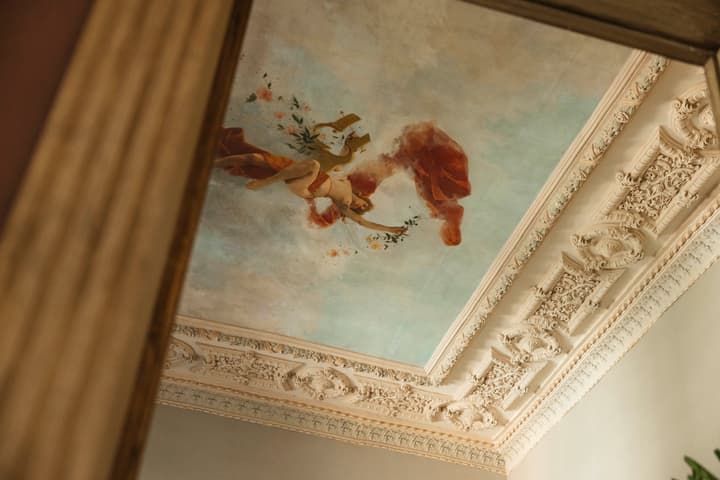 A fresco painting of a figure holding flowers adorns an ornate, intricately detailed ceiling inside a classical, elegant interior with decorative molding.