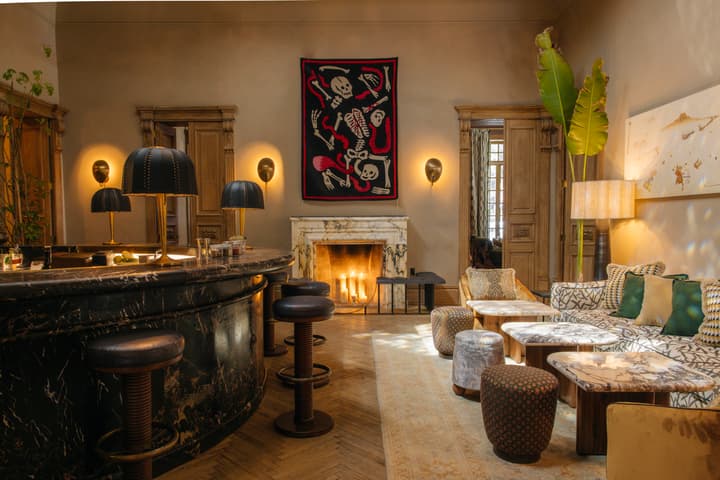 A cozy, dimly lit room featuring a marble bar counter with bar stools, a plush seating area with patterned cushions and tables, and a lit fireplace beneath an eclectic artwork of skeletons.