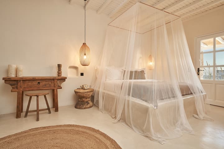 A four-poster bed with sheer white drapes is illuminated by hanging pendant lamps in a minimalistic bedroom with light-colored walls, wooden furniture, a round woven rug, and a large window overlooking a bright, outdoor area.