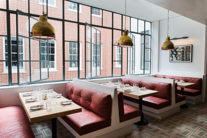 Red leather booths surround tables set with white napkins and glassware in a modern, brightly lit restaurant. Large windows overlook a brick building courtyard.