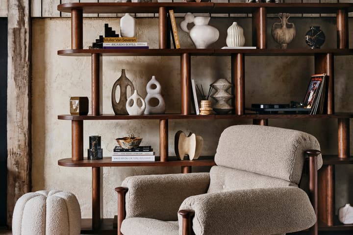 A plush armchair with wooden frame sits on a patterned rug in front of a wooden shelf filled with decorative sculptures, books, and vases, in a rustic room.