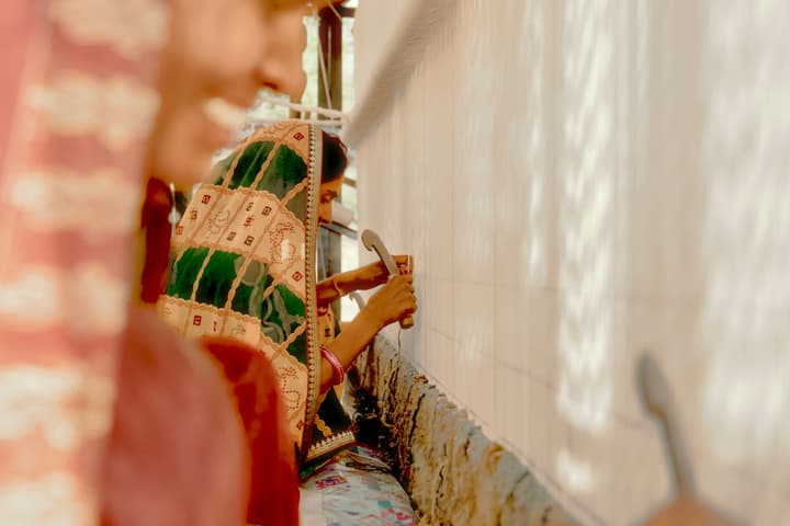 A woman in a patterned sari is working with a tool against a vertical woven fabric in an indoor setting with bright lighting. Another woman in the foreground appears out of focus.