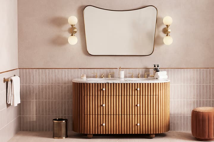 Wooden vanity with marble top, holding toiletries and rolled towels, against a tiled wall, below a large abstract-shaped mirror flanked by two globe lights in a modern bathroom.