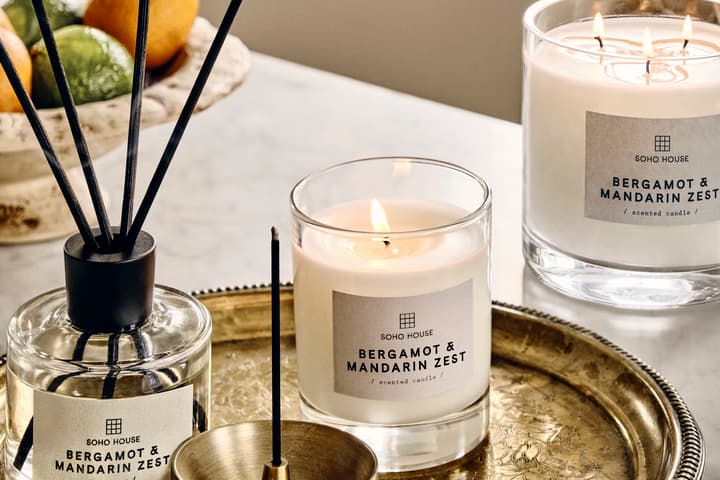 Lit and unlit Soho House “Bergamot & Mandarin Zest” candles and a diffuser sit on a decorative brass tray on a marble table, with a fruit bowl in the background.
