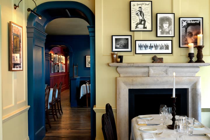 A dining table with a lit candle is placed near a stone fireplace adorned with framed pictures, set in a warmly lit restaurant with blue and yellow walls and a gallery-lined hallway.