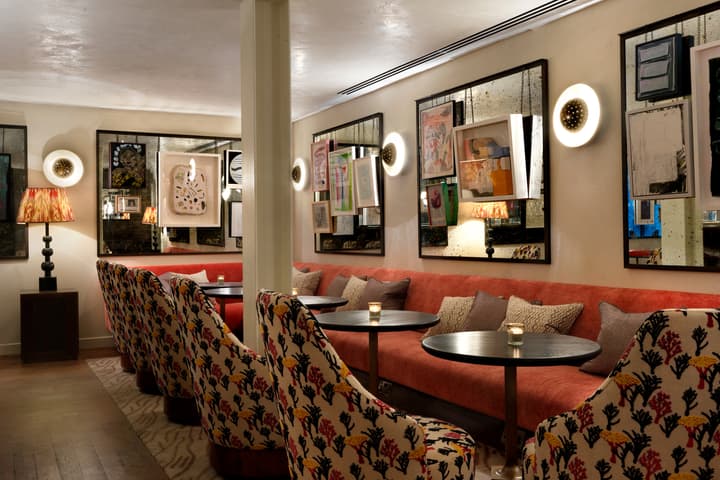 Plush, patterned chairs surround small round tables in a warmly lit, art-filled lounge, featuring a long red sofa, pillows, framed artwork, wall sconces, and a decorative table lamp.