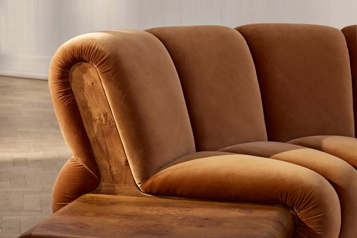 A plush, curved brown couch with wooden armrests is situated on a wooden floor in a white, minimally-decorated room.