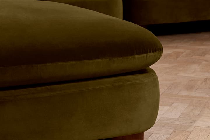 A green velvet sofa with rounded wooden legs is positioned on a polished herringbone-patterned wooden floor in an indoor setting.
