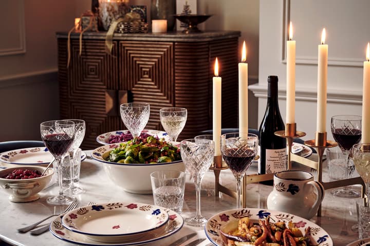 A dinner table set with patterned plates, wine glasses, and bowls of food, surrounded by lit candles; in the background is a cabinet with decoratively arranged candles and flowers.