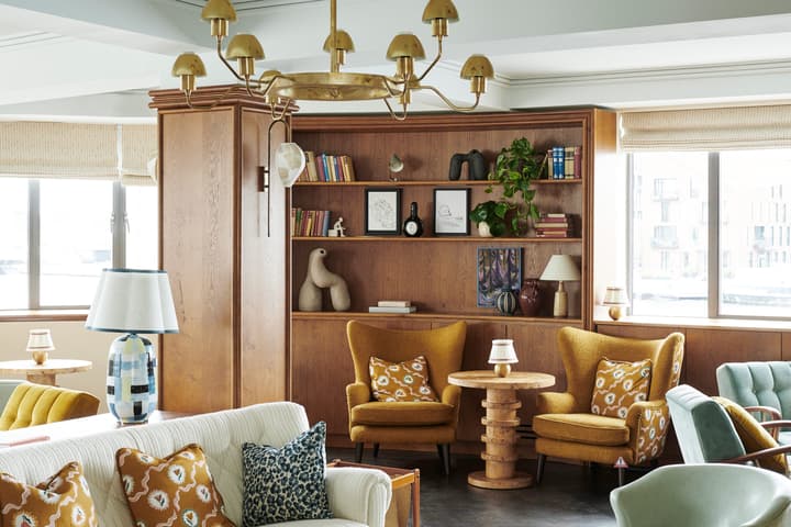 A cozy living room features a white sofa adorned with floral pillows, facing a geometric coffee table on a blue patterned rug. Background includes bookshelves, armchairs, and large windows.