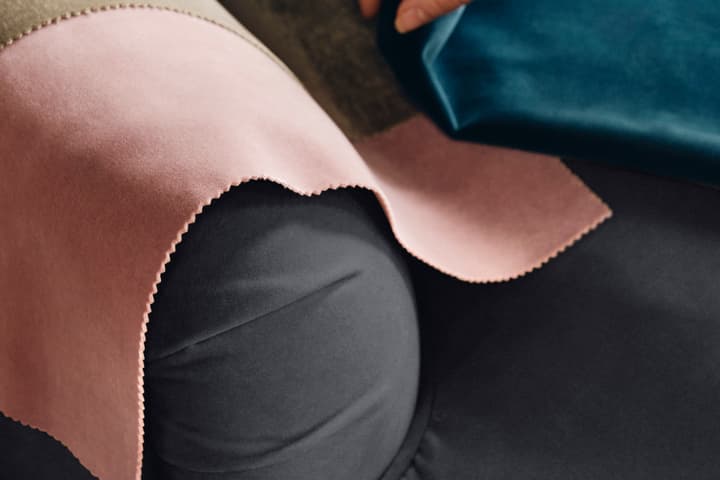 A hand places various fabric swatches, including pink, beige, and blue, onto a dark-colored upholstered armrest in a dimly lit, indoor setting.