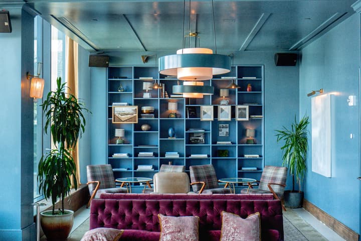 Luxurious, colorful lounge with vibrant patterned couches and armchairs, lit by hanging lamps. The space includes large potted plants and a backdrop of minimalist shelving with assorted decorations.