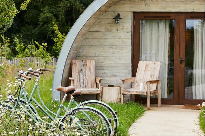 Curved wooden cabin with glass doors, two wooden chairs outside, two bicycles parked nearby, all amidst greenery and wildflowers, surrounded by trees in a serene nature setting.