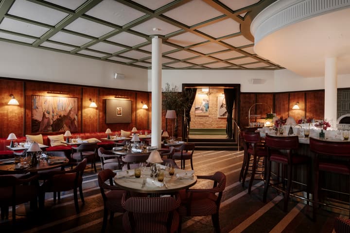 Restaurant with round tables and striped chairs, garnished with tableware and small lamps, alongside a bar counter with high stools, situated in a warmly lit, art-adorned, wood-paneled space.