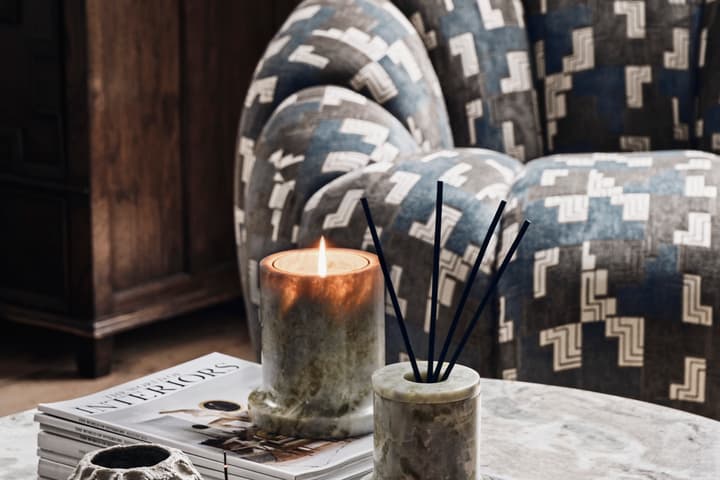 A lit candle sits on stacked magazines alongside incense and decor items on a marble table. In the background, there is a patterned, upholstered chair, and a dark wooden cabinet.