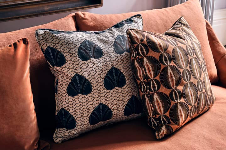 Decorative pillows with intricate patterns rest on an orange-brown couch, next to a window or wall and radiator, in a cozy living room.