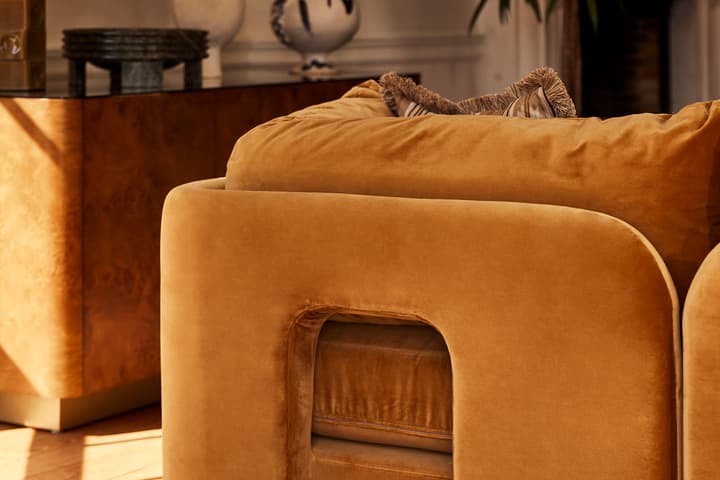 A plush, mustard-colored armchair with a rectangular cut-out in a stylish, sunlit living room, surrounded by ceramic vases, a side table with a lamp, and a potted plant.