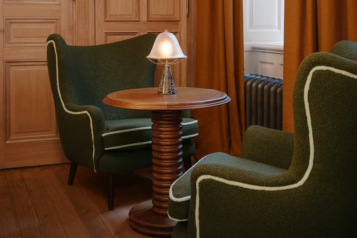 Green upholstered chairs face a round wooden table centered with a lit decorative lamp, surrounded by wood-paneled walls, orange curtains, and a radiator beneath a window.