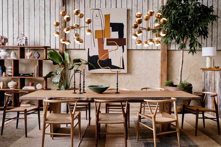 A wooden dining table with chairs is adorned with black candlesticks and a green bowl, set in a room with plants, shelves displaying decor items, and abstract art on the wall.