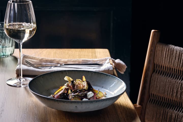 A ceramic bowl with a gourmet salad sits on a wooden table beside a wine glass, cutlery, and a folded napkin; a chair with a woven seat is pushed nearby.