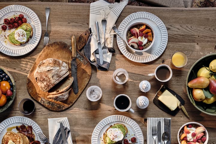 Plates with avocado toast, granola bowls, and pancakes surround a loaf of bread on a wooden table, with cutlery, fruit, coffee, juice, and butter scattered among them.