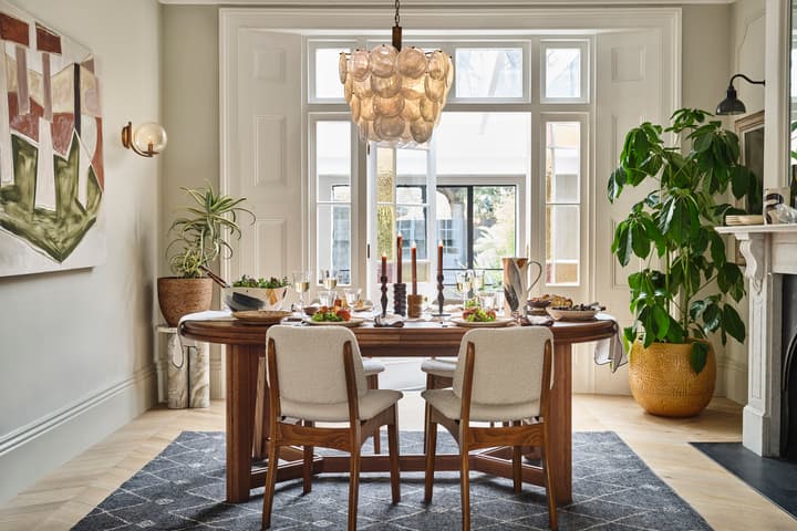 A wooden dining table set for a meal, featuring plates, glasses, and candles, surrounded by cushioned chairs in an elegant room with large windows, a plant, and modern artwork.