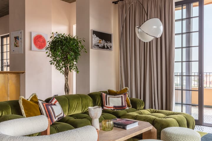 A modern living room with a green velvet sectional sofa, abstract-patterned pillows, and a wooden coffee table displaying books; a white armchair, green ottomans, floor lamp, and wall art add detail.