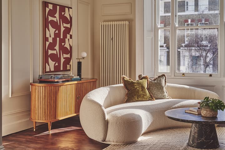 A curvy white sofa with patterned pillows sits in a well-lit living room, facing a round black marble coffee table, with a wooden sideboard and abstract framed artwork nearby.
