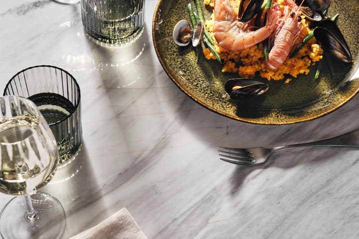 A green ceramic plate with seafood paella, surrounded by three glasses of white wine and a fork, all placed on a marble table with a linen napkin.