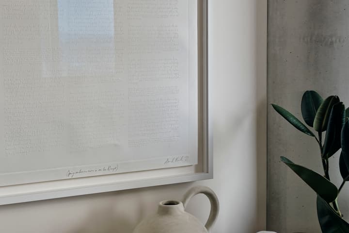 A framed document with handwritten text hanging on a white wall, next to a potted plant and a gray ceramic jug on a table.