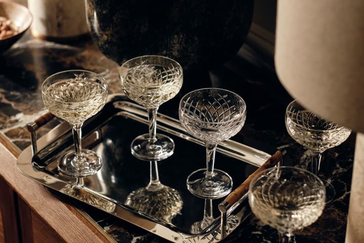 Five crystal champagne coupes filled with a drink sit on a polished, silver tray on a marble-topped table, flanked by decorative objects and under soft lighting.