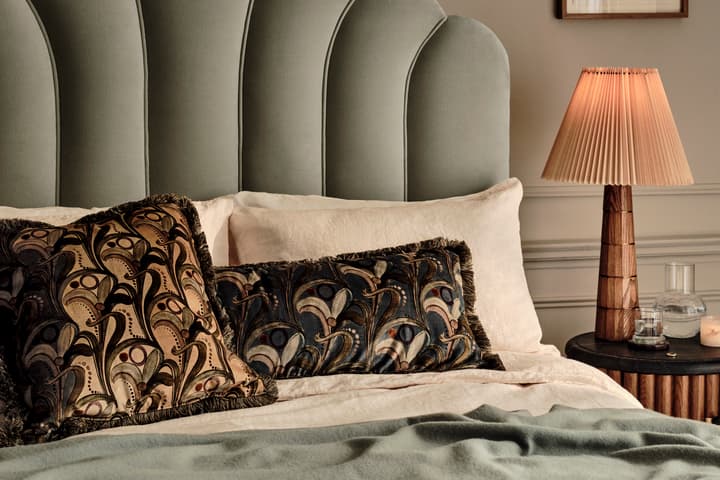 Bed with patterned pillows resting against a grey upholstered headboard, next to a wooden nightstand with a pleated lampshade, glass of water, and candle, situated in a beige-colored room.