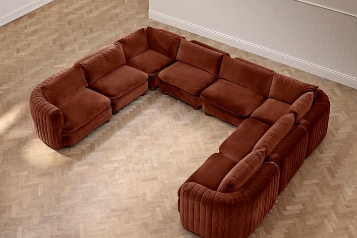 A large, U-shaped, brown velvet sectional sofa with plush cushions sits on a light brown herringbone-patterned wooden floor in a minimalistic room with white walls.