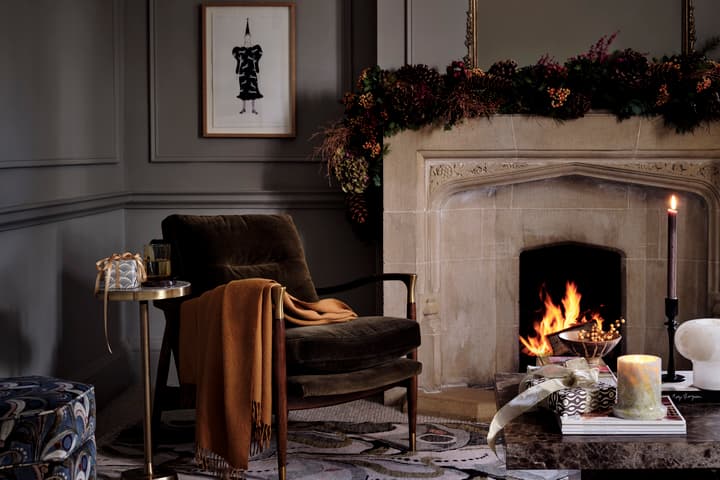 A cushioned armchair with an orange blanket draped over it, beside a small table holding a gift and glass, faces a decorated stone fireplace with a roaring fire in a dimly lit room.