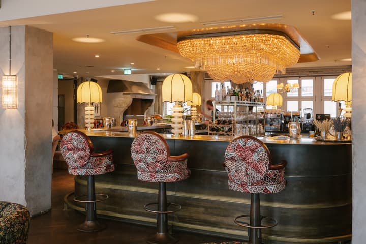 Ornate bar counter with red floral cushioned stools beneath a large chandelier, attended by a bartender in a warmly lit, modern space with beige walls and large windows.